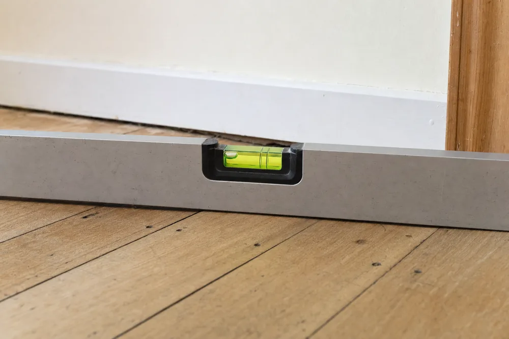 A spirit level lying on a timber floor with its bubble at one end of the vial, beside a skirting board with a dark gap under it — house levelling, Newcastle. Illustrative example.